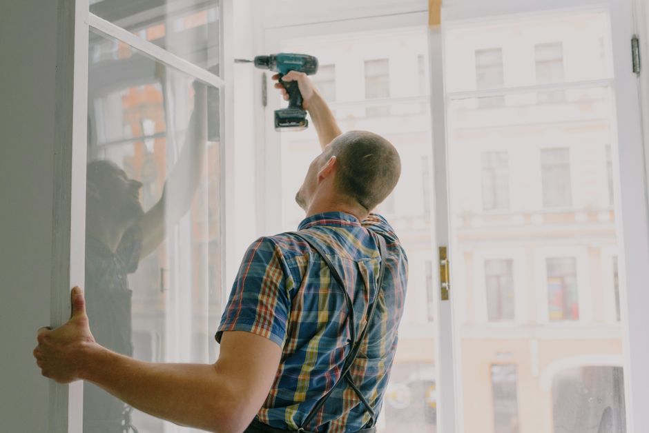 Worker in a checkered shirt drilling a window frame into a wall opening.